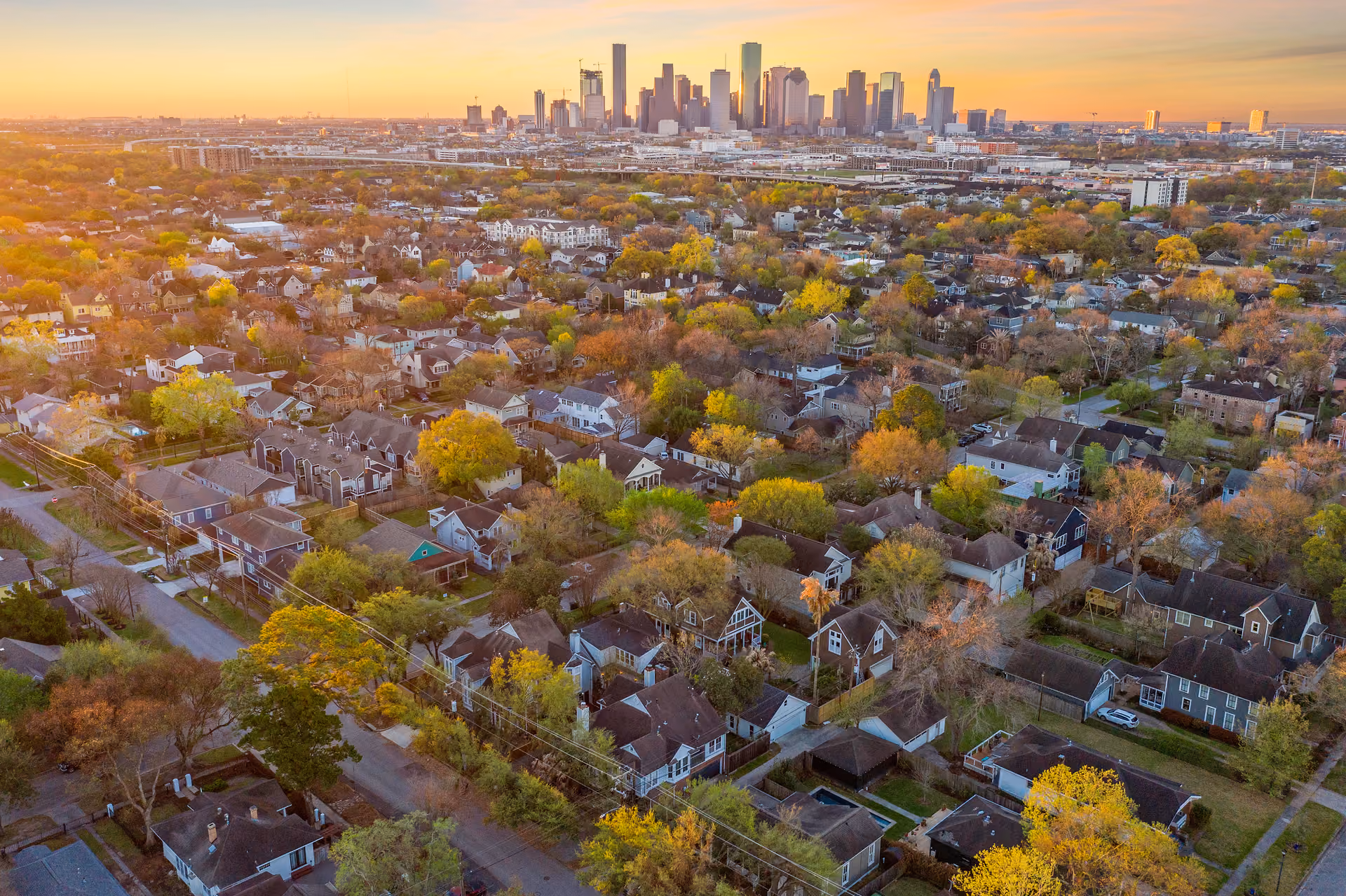 Aerial view over Houston Heights at golden hour: blocks of single-family houses under mature trees, with the downtown Houston skyline on the horizon.